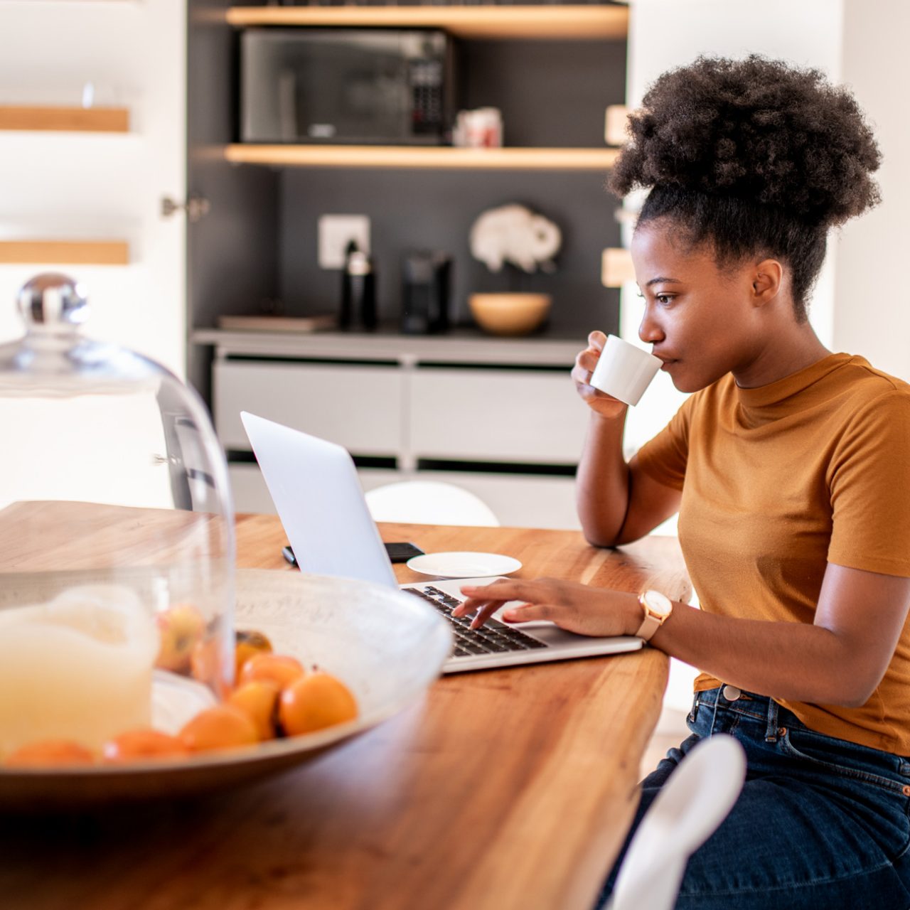 Woman seated at table in kitchen tapping key on laptop while drinking from cup