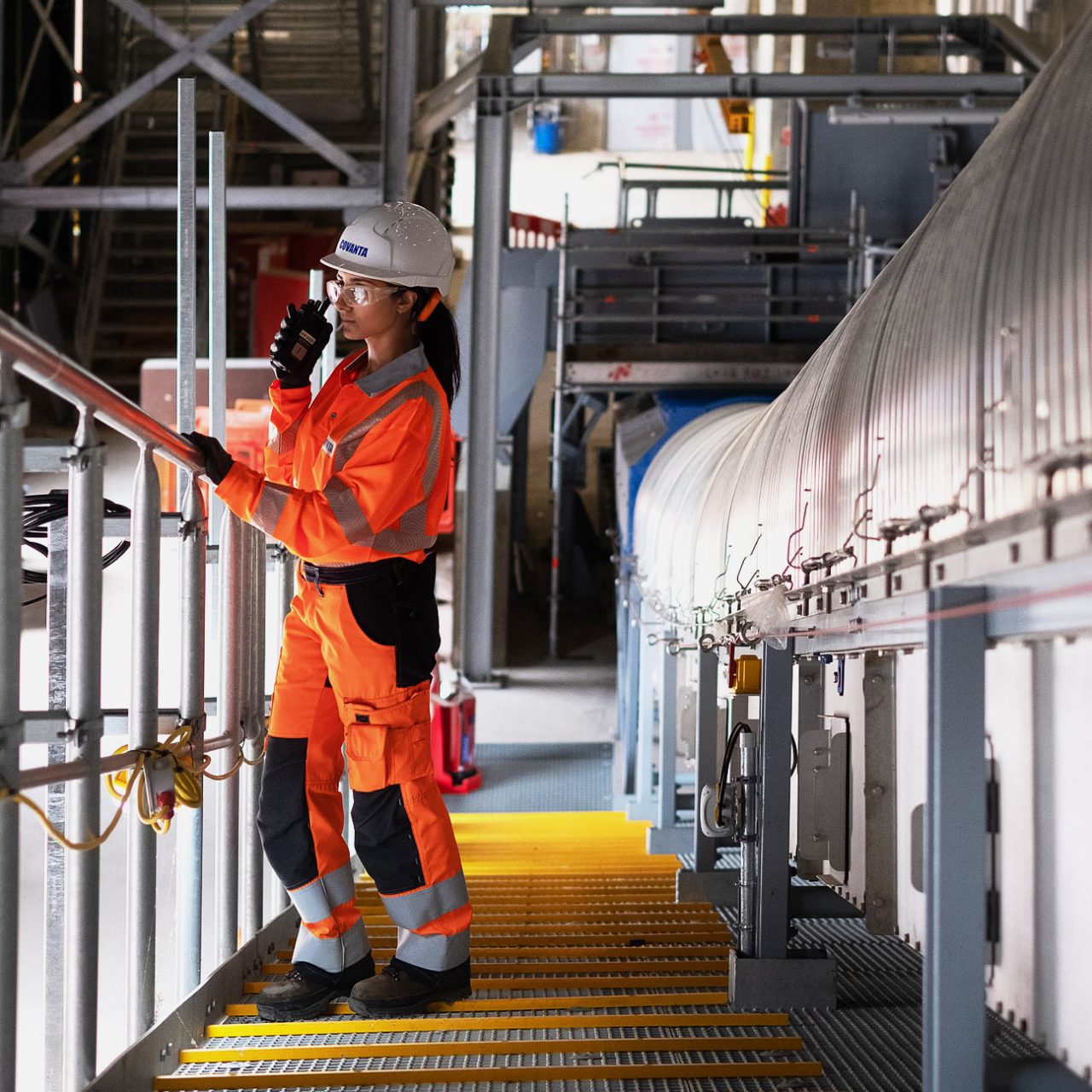 Woman in white hard hat and orange boiler suit leaning on metal railing using handheld receiver