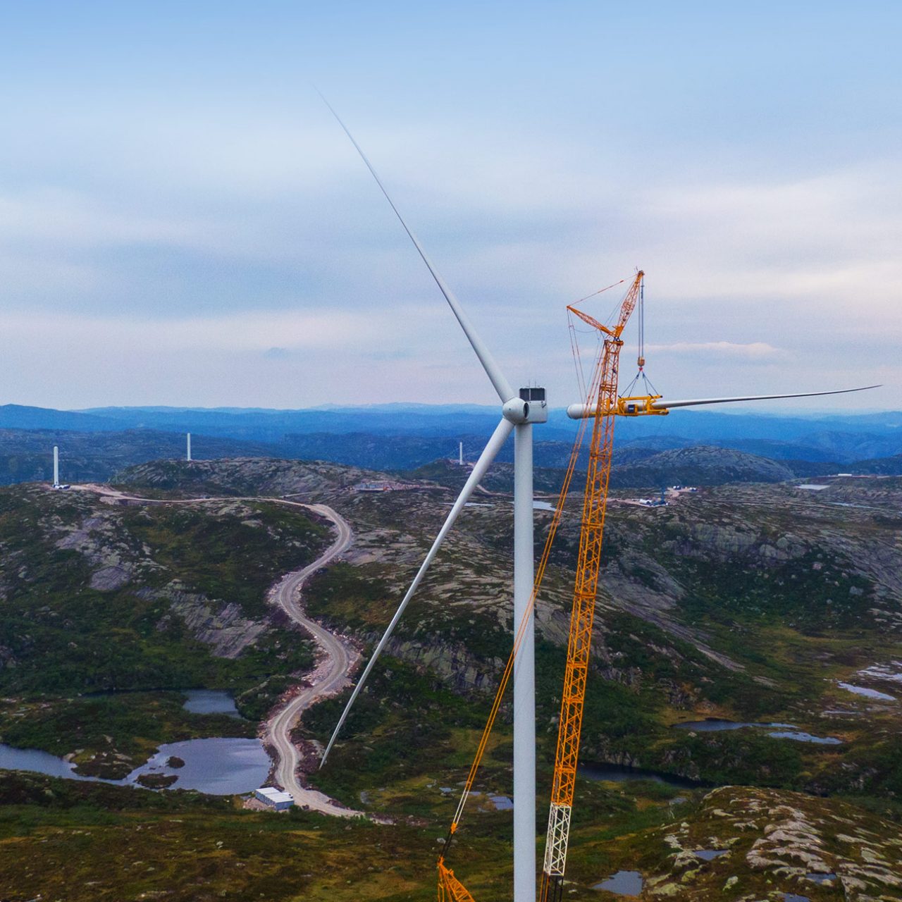 Yellow crane attaching wind turbine blade against hills and blue sky