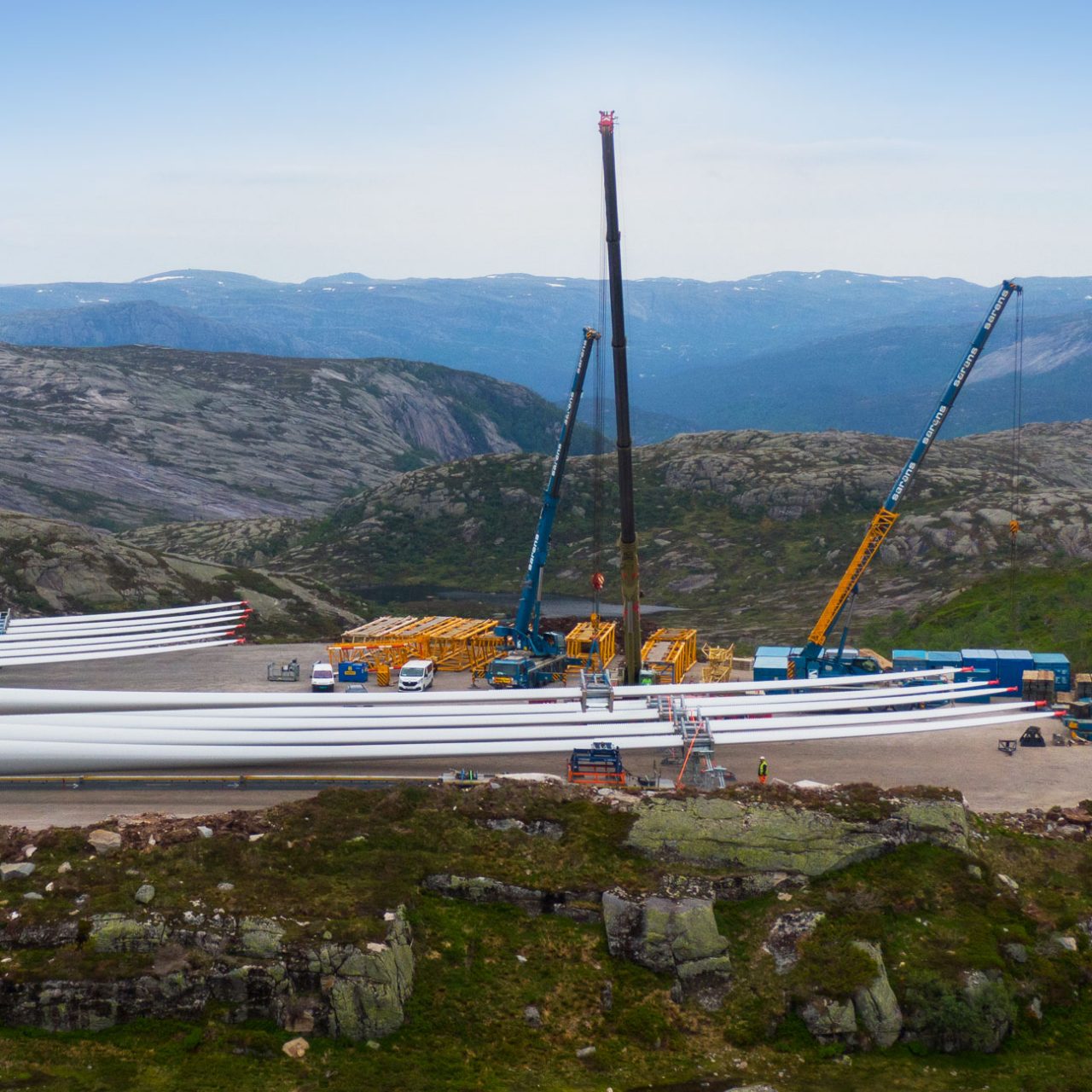 Wind turbine blades lined up on ground with cranes and hills in background