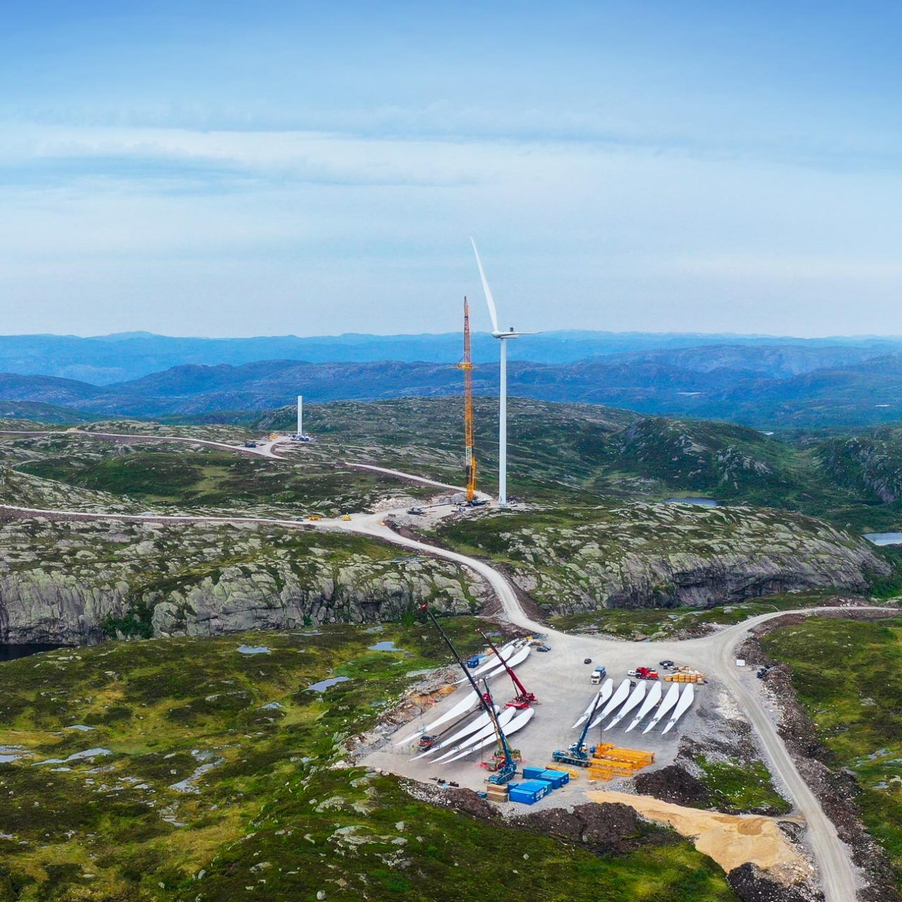 Wind turbines blades lined up on ground in foreground; wind turbine and crane in background with blue sky