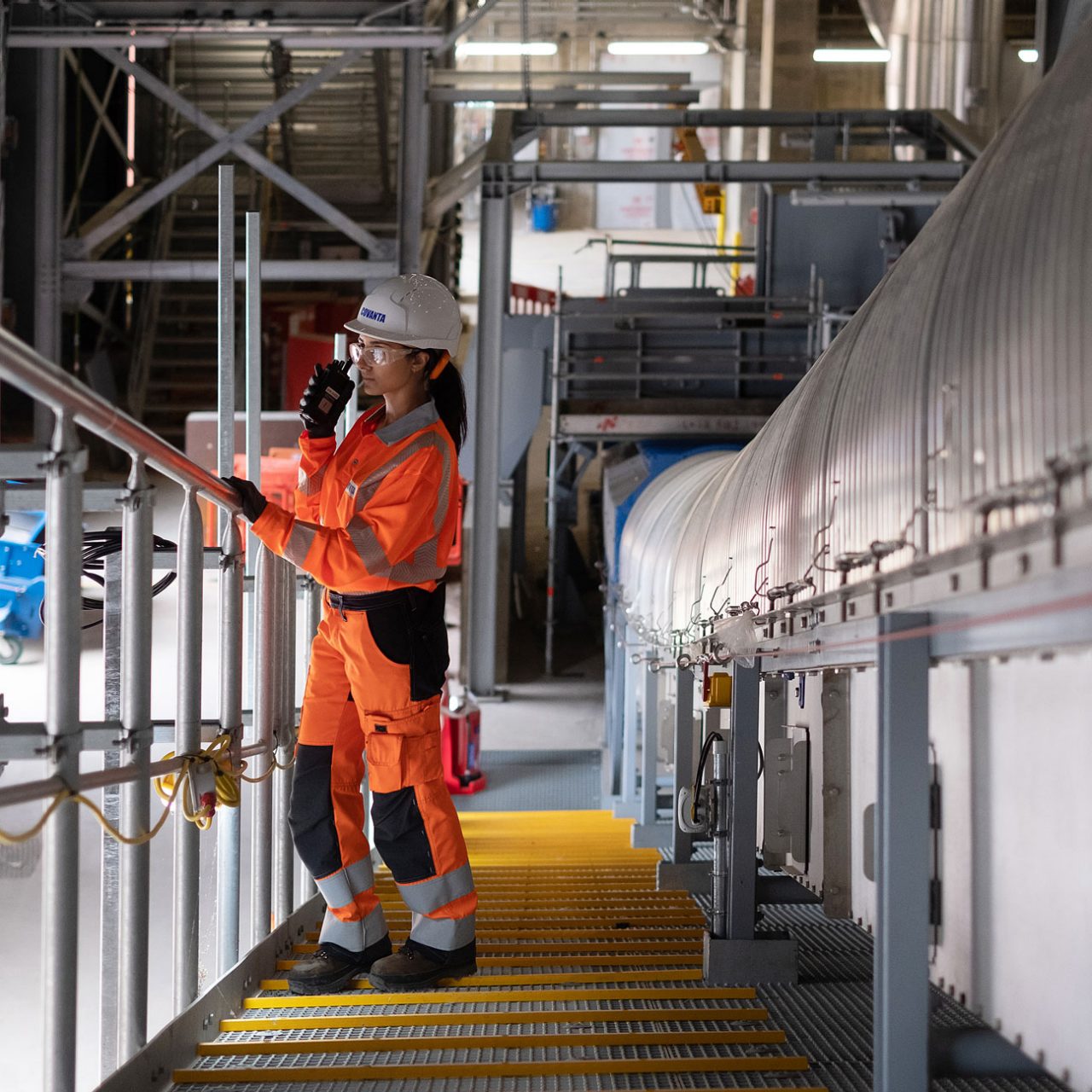 Woman in white hard hat and orange boiler suit leaning on metal railing using handheld receiver